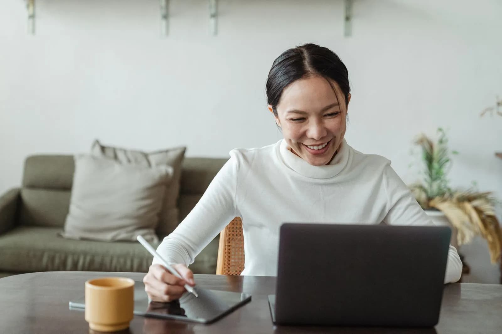 Young smiling Asian female communicating with colleagues on laptop and making notes on tablet with stylus while working remotely at home