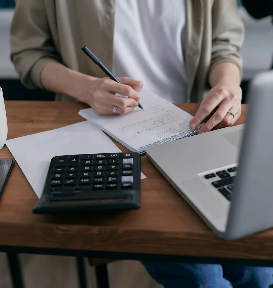 A woman writes financial calculations in a notebook, using a calculator and laptop at a wooden desk